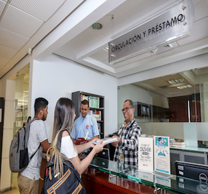 esta es una imagen de unos estudiantes recibiendo un libro en el área en el escritorio de Solicitud y Préstamo de la biblioteca